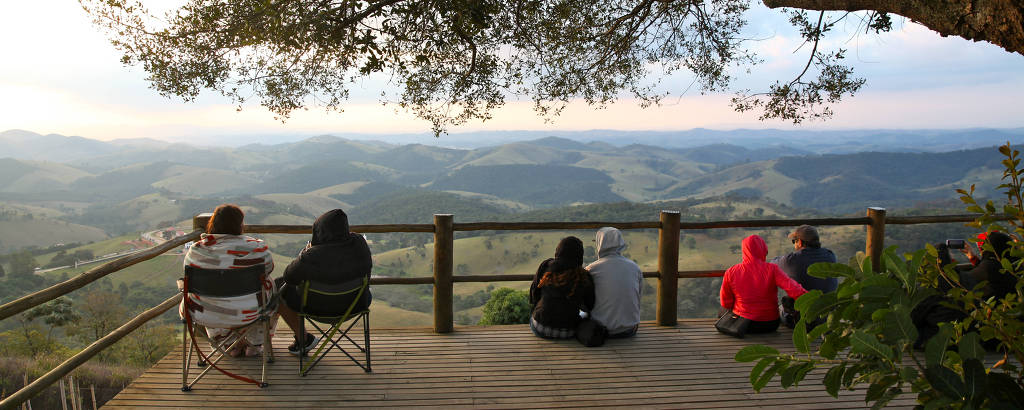 A imagem mostra um grupo de pessoas sentadas em um deck de madeira, observando uma paisagem montanhosa ao fundo. Há uma grande árvore à esquerda, e as pessoas estão vestidas com roupas de frio, algumas com capuzes. A cena é tranquila, com montanhas e vales visíveis sob um céu claro.