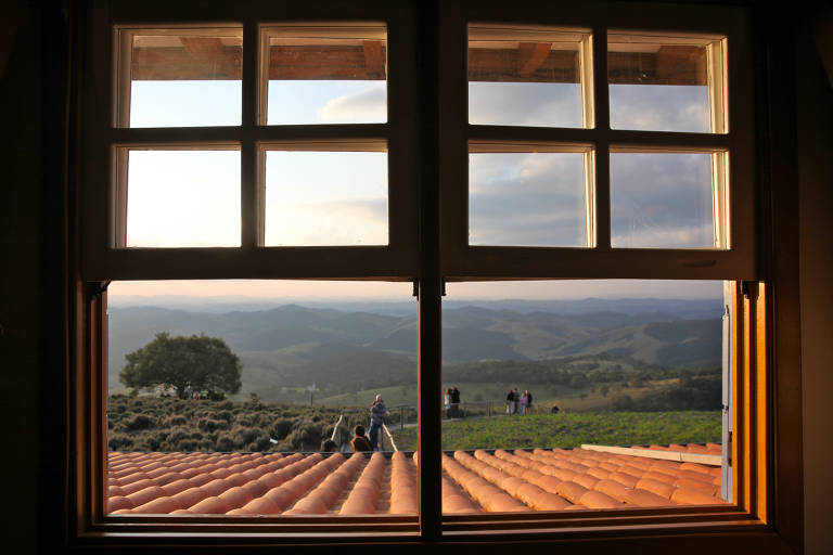 A imagem mostra uma janela com quatro painéis de vidro, através da qual se vê uma paisagem montanhosa ao fundo. O céu está parcialmente nublado e a luz do sol ilumina a cena, criando um efeito suave. Na parte inferior, há um telhado de cerâmica laranja e, ao fundo, algumas pessoas estão em pé, observando a vista. Árvores e colinas verdes são visíveis na paisagem.