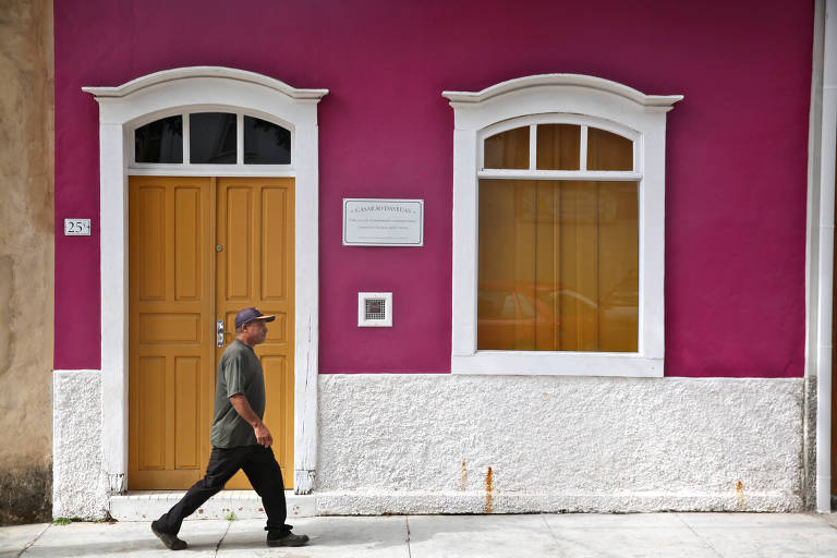 Um homem caminha em frente a uma parede rosa com uma porta amarela e janelas brancas. A porta possui um número 25 e há uma placa ao lado que diz 'CASARÃO DAS LÚLIAS'. A janela à direita tem cortinas amarelas.