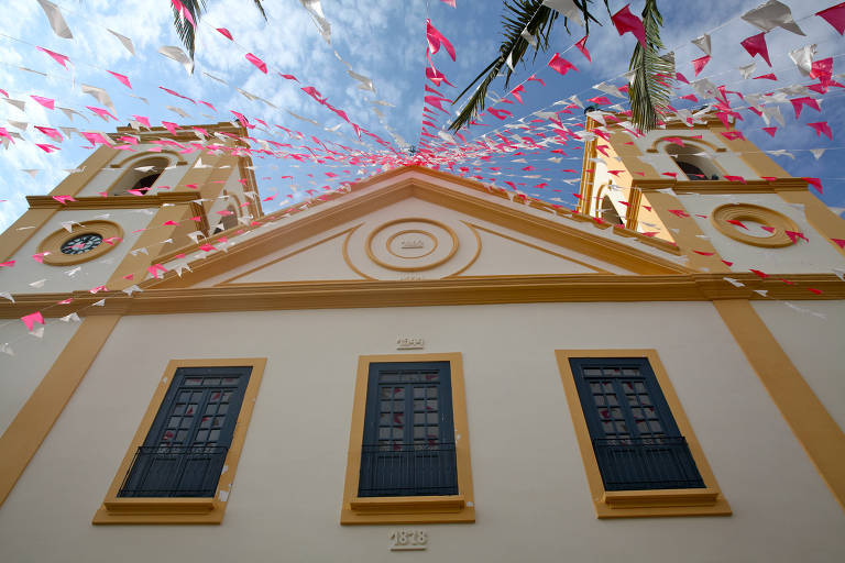 A imagem mostra a fachada de uma igreja vista de baixo para cima, com duas torres laterais. A igreja é predominantemente branca com detalhes em amarelo. Há bandeirinhas em cores rosa e branca penduradas no ar, criando um efeito festivo. O céu está parcialmente nublado, e a data de construção da igreja é visível nas paredes, com os anos 1824, 1844 e 1878 destacados.