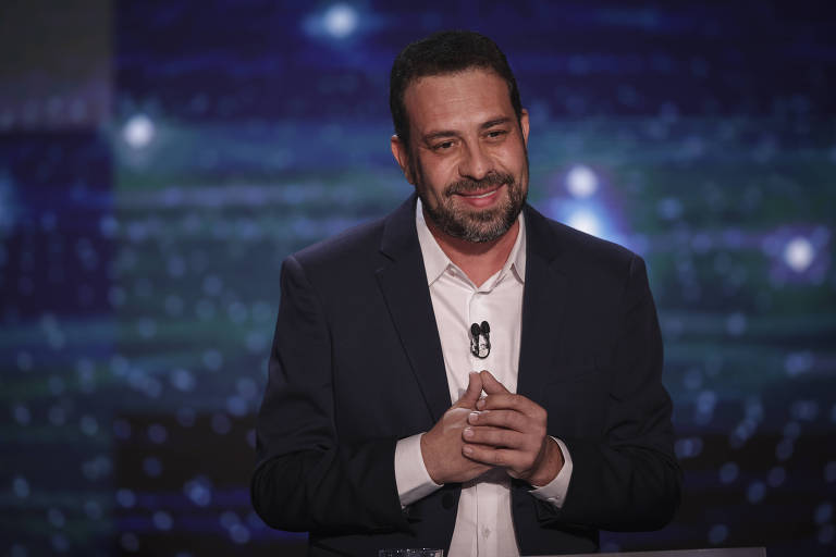 Um homem está em um palco， sorrindo e com as mãos unidas na frente do corpo. Ele usa um paletó escuro e uma camisa branca. O fundo é iluminado com luzes azuis e brancas， criando um efeito de brilho.