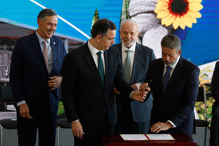 Rodrigo Pacheco passa uma caneta para Arthur Lira, sendo que os dois seguram o objeto ao mesmo tempo, sob os olhares de Lula e Barroso, todos em frente a um painel com o desenho de uma flor amarela