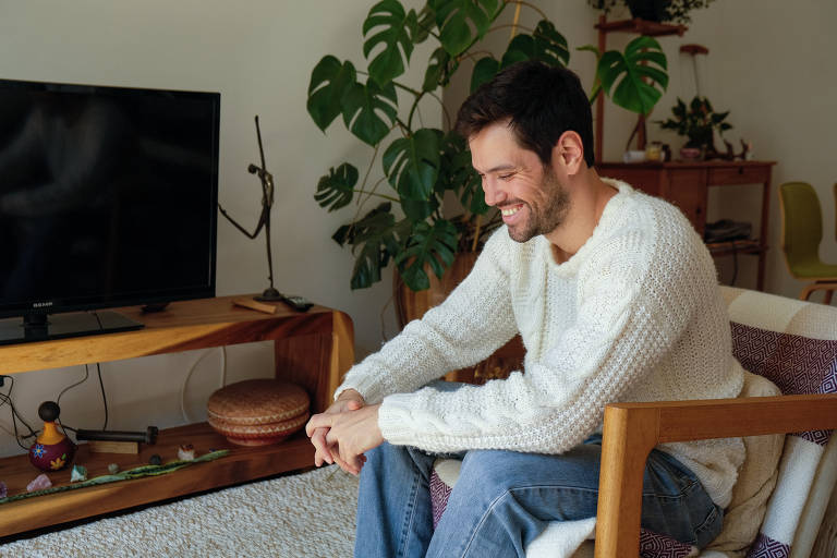 Uma visão lateral do homem sentado, ainda sorrindo. Ele está inclinado levemente para a frente, com os braços repousando nos joelhos, enquanto mantém a mesma expressão amigável e descontraída.