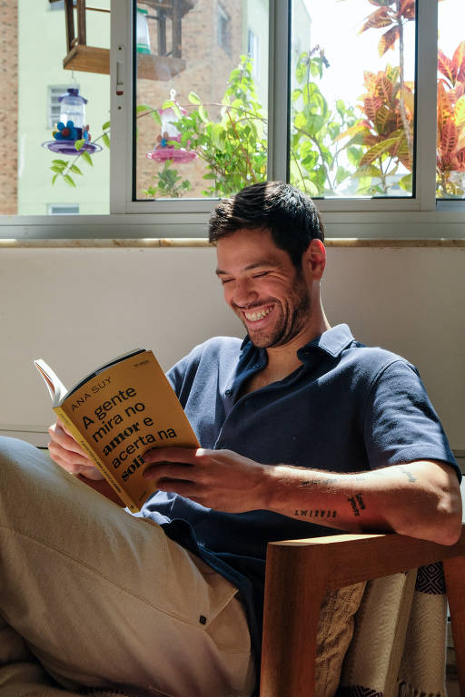 Um homem sentado em uma cadeira perto de uma janela, rindo enquanto lê um livro intitulado "A gente mira no amor e acerta na solidão", de Ana Suy. Ele está usando uma camisa polo azul, e ao fundo, plantas e uma janela deixam a luz do sol entrar.