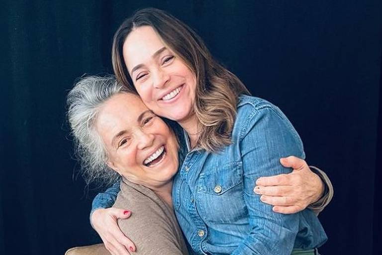 Em foto colorida, duas mulheres posam juntas sentadas em um sofá