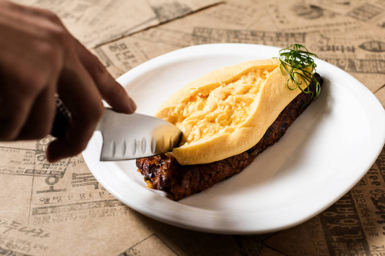 A imagem mostra um prato com uma omelete amarela em cima de uma porção de arroz de costela. Uma mão segurando uma faca está prestes a cortar a omelete. O fundo é composto por papel com texto impresso.