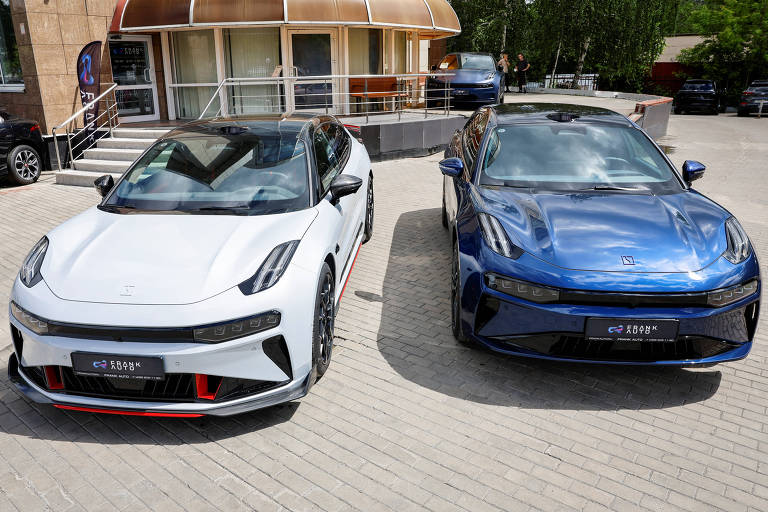 A imagem mostra dois carros estacionados em uma área externa. O carro à esquerda é branco com detalhes em preto, enquanto o carro à direita é azul. Ambos estão em frente a um edifício com uma fachada de vidro e uma cobertura arredondada. O chão é de pedras claras e há outros veículos ao fundo.