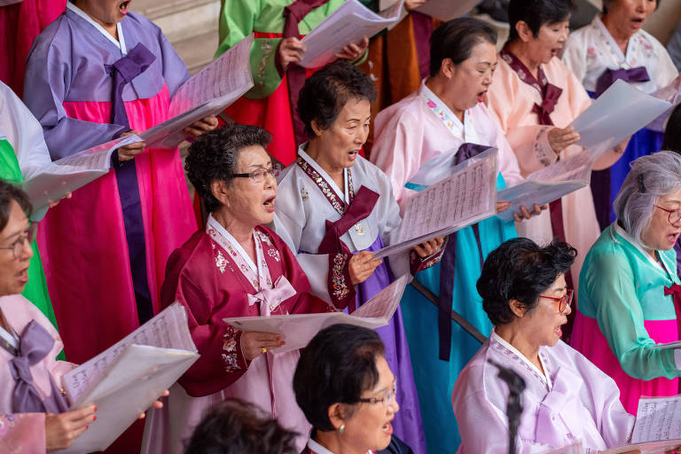 Um grupo de mulheres vestindo hanboks coloridos, cantando em um coral. Elas estão segurando partituras e expressando alegria enquanto cantam. As roupas variam em cores, incluindo rosa, azul, verde e roxo, refletindo a tradição cultural. O ambiente parece ser um espaço público ou um evento cultural.