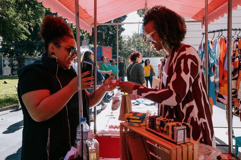 A imagem mostra um mercado ao ar livre com um estande coberto por uma tenda vermelha. Duas mulheres estão interagindo: uma está segurando um celular e a outra está entregando um item. Ao fundo, há várias pessoas, algumas vestindo camisetas coloridas, e um suporte com roupas penduradas. O ambiente é ensolarado, com árvores ao fundo.