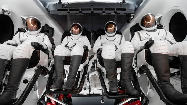 A imagem mostra quatro astronautas sentados em cadeiras dentro de uma nave espacial. Eles estão vestidos com trajes espaciais brancos e capacetes com visores escuros. O interior da nave é moderno, com paredes brancas e detalhes em preto, e há janelas circulares ao fundo.