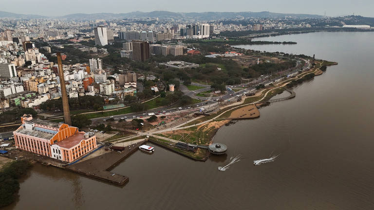 A imagem mostra uma vista aérea de uma cidade localizada à beira de um rio. À esquerda, há um edifício de cor laranja com uma chaminé alta. O rio é de cor marrom e há pequenas embarcações navegando. Ao fundo, a cidade é composta por vários prédios e montanhas ao longe, com uma vegetação ao longo da margem do rio.