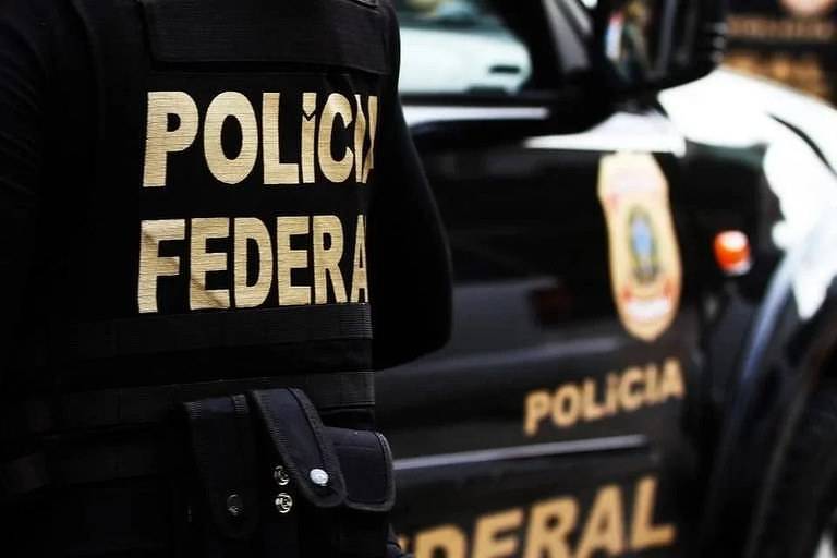 A imagem mostra um agente da Polícia Federal com um colete preto, onde está escrito 'POLÍCIA FEDERAL' em letras douradas. Ao fundo, é visível um veículo da Polícia Federal, também com a inscrição 'POLÍCIA' e o emblema da corporação.