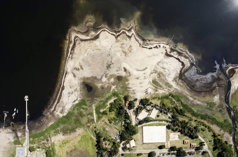 Imagem aérea de uma área costeira, mostrando uma combinação de terra e água. A parte inferior da imagem apresenta uma faixa de terra clara, possivelmente areia ou lama, com contornos irregulares. À direita, há uma área verde com vegetação e estruturas construídas, enquanto à esquerda, a água é visível, com uma transição entre a terra e o lago. A imagem captura a interação entre o ambiente terrestre e aquático.