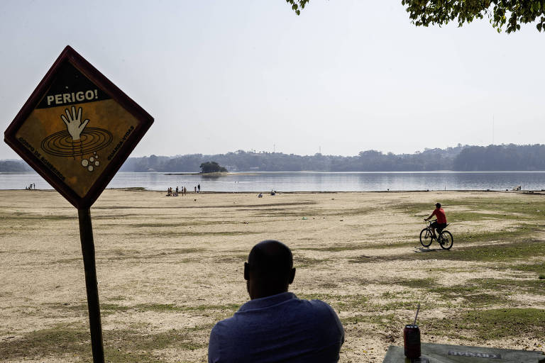 A imagem mostra uma área de praia com um homem sentado de costas, observando o cenário. À sua frente, há um sinal de advertência que diz 'PERIGO'. No fundo, é possível ver um lago e algumas pessoas, incluindo uma que está andando de bicicleta na areia. A vegetação ao redor é escassa e o céu está claro.