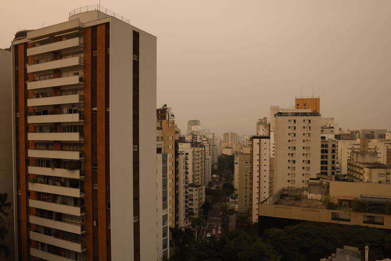 A imagem mostra uma vista de um bairro urbano com vários edifícios altos. O céu está nublado, com uma tonalidade amarelada, sugerindo poluição ou fumaça. Os prédios são de diferentes estilos arquitetônicos, com predominância de cores claras e algumas faixas de madeira. A rua entre os edifícios é visível, mas parece vazia.