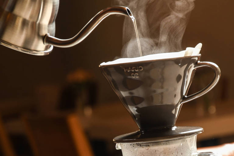 A imagem mostra um processo de preparo de café, onde um bule de metal está vertendo água quente sobre um filtro de café preto. A água quente gera vapor, que é visível na imagem. Abaixo do filtro, há um recipiente transparente contendo café coado. O fundo da imagem apresenta um ambiente de café, com mesas e cadeiras desfocadas.