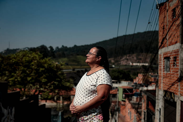 Uma mulher está em pé, olhando para o horizonte em uma comunidade. Ela usa uma blusa branca com bolinhas e uma saia colorida. Ao fundo, há casas de tijolos e uma vegetação montanhosa. O céu está claro e azul, e há fios elétricos visíveis na imagem.