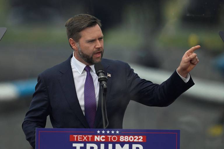 Homem branco, de barba, usando terno e gravata escuros, e camisa branca, fala atrás de pódio com a palavra "Trump"