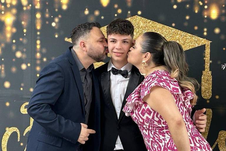 Em frente a um painel dourado, Fabi e o esposo Ronaldo beijam o filho ao centro. Ela, à direita, é uma mulher branca, com cabelo castanho claro preso. Usa um vestido branco e vermelho. Guilherme, ao centro, é um menino branco e alto. Ele veste smoking. À esquerda, Ronaldo usa terno azul marinho. Ele é branco, tem cabelo castanho curto e barba escura e também beija o rosto do filho.