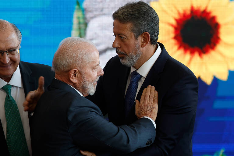 Lula, um homem branco de barba e cabelos ralos da mesma cor, coloca a mão sobre o peito de Lira, um homem branco, de barba e cabelo castanhos grisalhos. Os dois vestem terno e gravata escuros e ao fundo, ao lado deles, está o Alckmin, um homem branco, calvo, com terno e gravata verde. No telão, uma grande flor com pétalas amarelas 