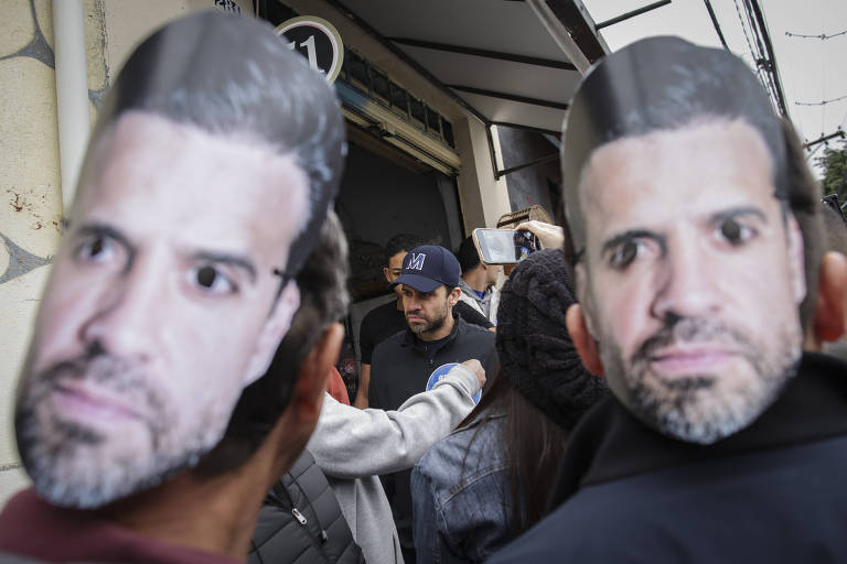 A imagem mostra um grupo de pessoas em uma rua, algumas delas usando máscaras de papel que representam o rosto de um homem. Ao fundo, há uma pessoa que parece estar sendo filmada ou fotografada. O ambiente é urbano, com uma entrada de loja visível.