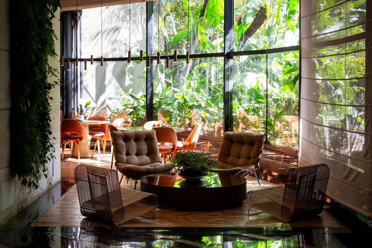 A imagem mostra um ambiente do hotel Renaissance iluminado por luz natural, com grandes janelas que oferecem vista para um ambiente verde e tropical. No centro, há uma mesa redonda com um vaso de plantas, cercada por poltronas de design moderno. Ao fundo, é possível ver uma área de refeições com cadeiras laranjas, também cercada por vegetação.