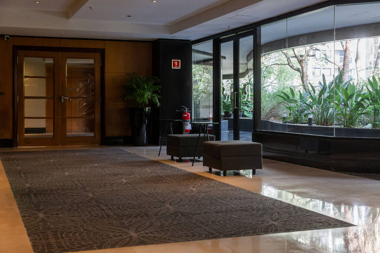 A imagem mostra o interior do lobby do Hotel Renaissance. À esquerda, há portas de vidro com moldura de madeira. No centro, um tapete grande cobre o chão, e há dois sofás pequenos em uma mesa de centro. À direita, grandes janelas permitem a entrada de luz natural e oferecem vista para plantas verdes do lado de fora.