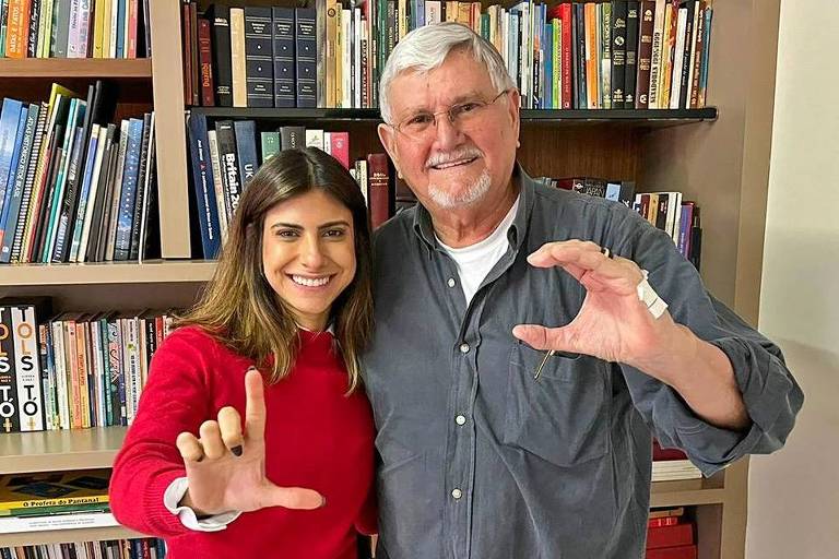 Zeca do PT faz um "C" as mãos. Ao seu lado, Camila Jara, de vermelho, faz um "L". Os dois estão num ambiente que parece uma biblioteca privada