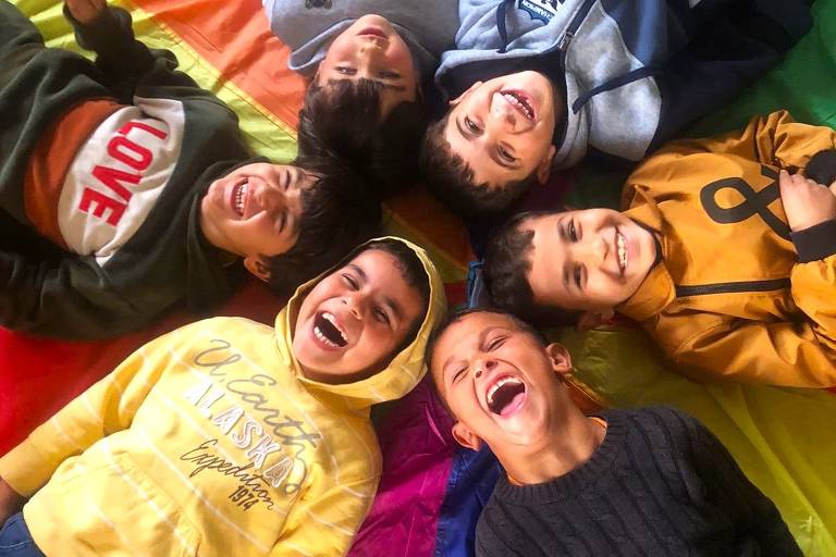 A imagem mostra seis crianças deitadas em um pano colorido, sorrindo e rindo. Elas estão dispostas em círculo, com os rostos voltados para cima. As crianças vestem roupas de diferentes cores, incluindo um moletom amarelo, um verde com a palavra 'LOVE', um cinza e um laranja. O pano em que estão deitados é uma bandeira colorida, com faixas em vermelho, laranja, amarelo, verde, azul e roxo.