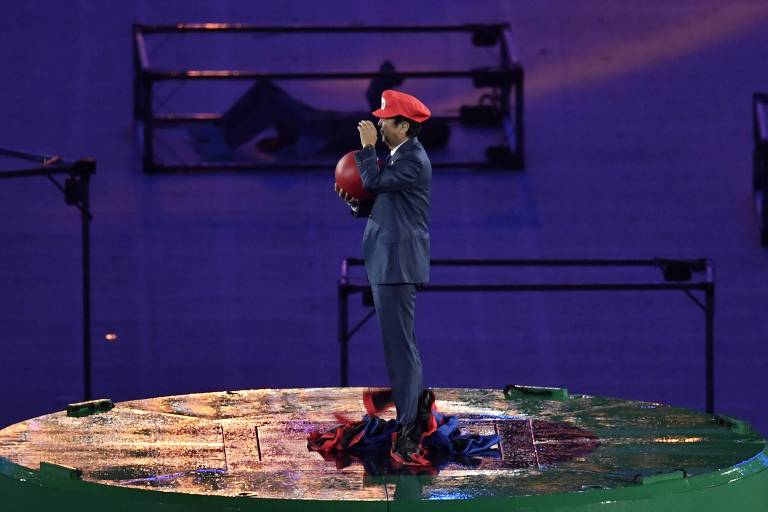 O então primeiro-ministro do Japão, Shinzo Abe, vestido de Mario, do jogo Mario Bros, na cerimônia de encerramento da Rio-2016. Abe veste um terno escuro e um chapéu vermelho está em pé em uma plataforma redonda, segurando uma bola vermelha. O ambiente é iluminado com luzes azuis e roxas, e ao fundo, há estruturas metálicas. A plataforma parece estar molhada, refletindo a luz.
