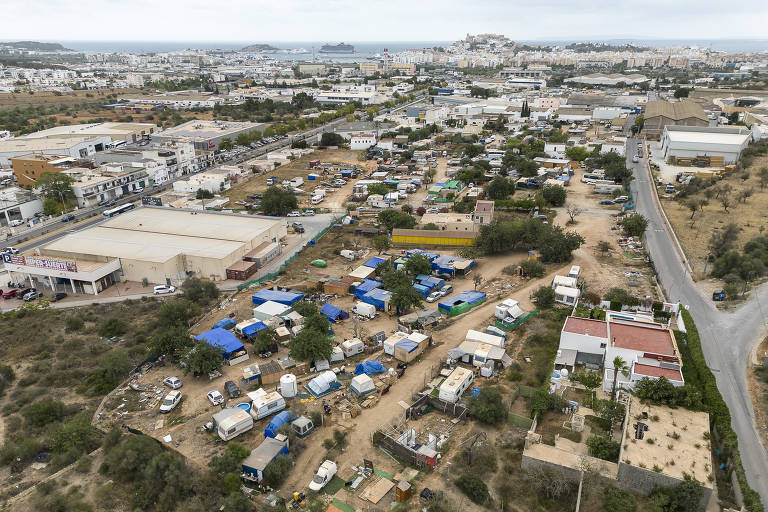 A imagem mostra uma vista aérea de uma área urbana com várias construções, incluindo armazéns e residências. No primeiro plano, há uma área com várias tendas e veículos, sugerindo uma ocupação informal. Ao fundo, pode-se ver uma cidade costeira com edifícios e um porto. O céu está nublado e há vegetação ao redor da área.