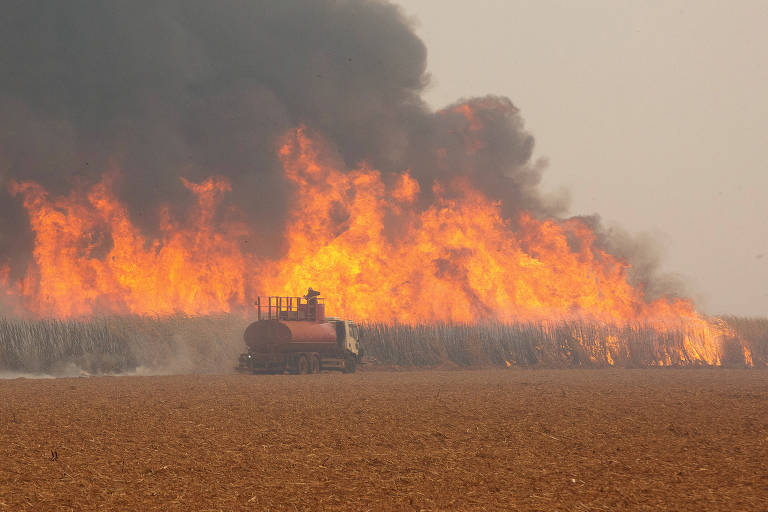 imagem mostra caminhão de bombeiros ao fundo, após campo com vegetação seca rasteira, e chamas que geram colunas de fumaça espessa, as chamas estão muito maiores do que o caminhão