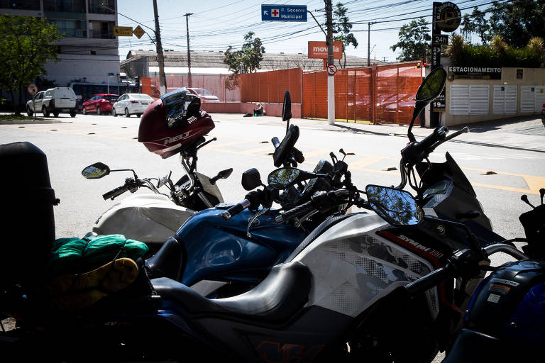 A imagem mostra várias motos estacionadas em uma calçada. Ao fundo， há uma rua com sinalização e uma obra cercada por uma tela laranja. O céu está claro e há alguns veículos visíveis na rua