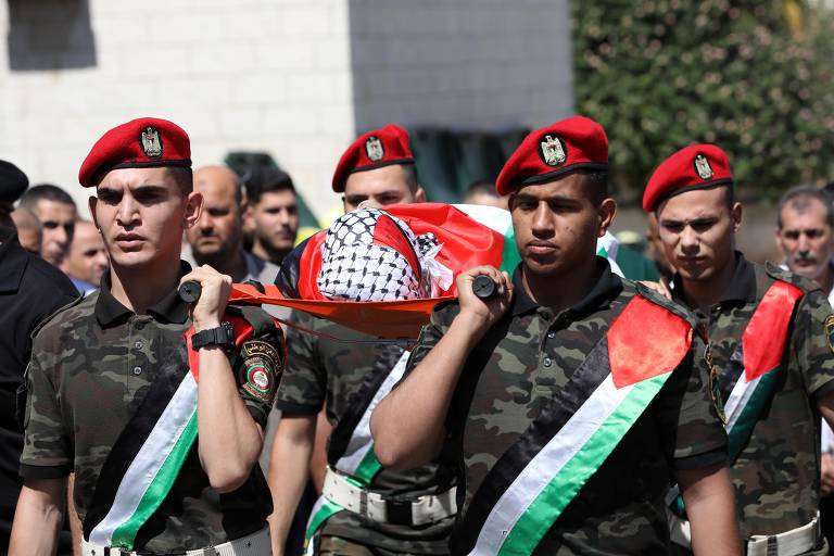 Um grupo de soldados em uniforme militar camuflado e boinas vermelhas carrega um caixão coberto com uma bandeira palestina. Os soldados estão alinhados e alguns deles usam faixas tricolores nas suas roupas. Ao fundo, há uma multidão e uma parede de pedra clara.