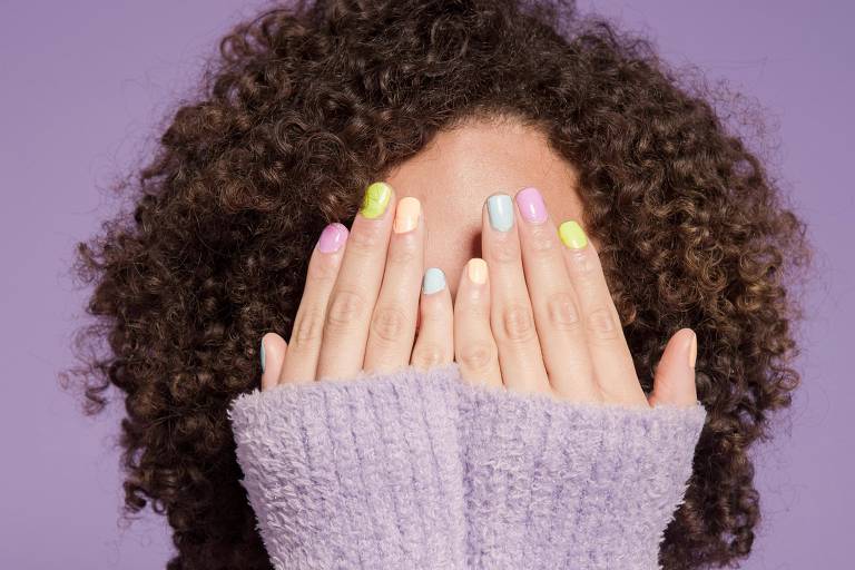 Uma pessoa com cabelo cacheado e volumoso está cobrindo o rosto com as mãos. Ela usa um suéter de cor lilás e suas unhas estão pintadas em várias cores pastel, incluindo rosa, azul, verde e amarelo. O fundo da imagem é de um tom lilás claro.