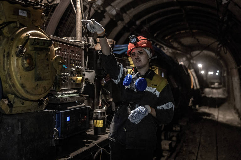 A imagem mostra uma trabalhadora em um túnel de mina, usando um capacete vermelho com uma lanterna na frente. Ela está vestindo um macacão de trabalho escuro com detalhes amarelos e brancos, e usa um protetor auricular azul. A mulher está apontando para um painel de controle ao seu lado, que possui vários botões e luzes. O ambiente é escuro, com iluminação suave ao fundo, e há equipamentos de mineração visíveis ao redor.