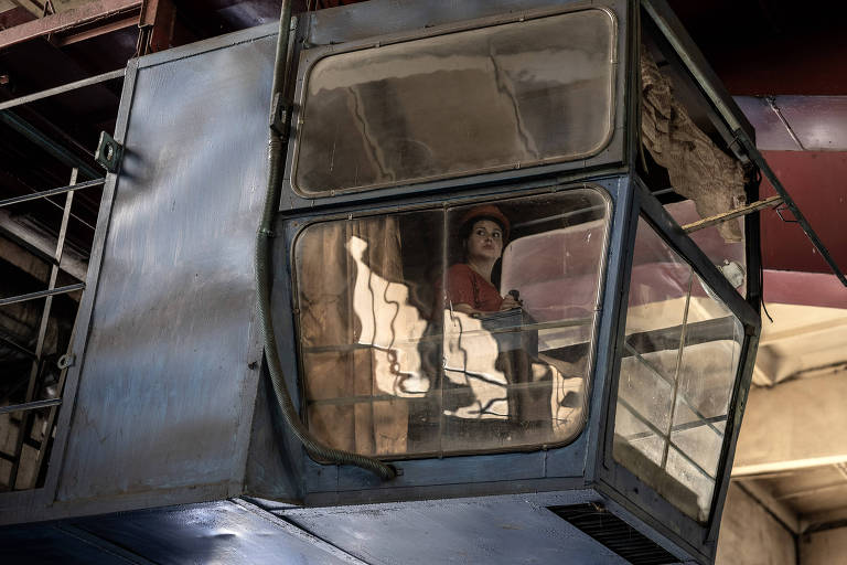 A imagem mostra uma mulher sentada dentro de uma cabine de guindaste, que está suspensa em um ambiente industrial. A cabine é de cor azul e possui janelas grandes, refletindo a luz do ambiente. A mulher usa uma blusa vermelha e um boné, olhando para fora da cabine. O fundo é composto por estruturas metálicas e um teto alto.