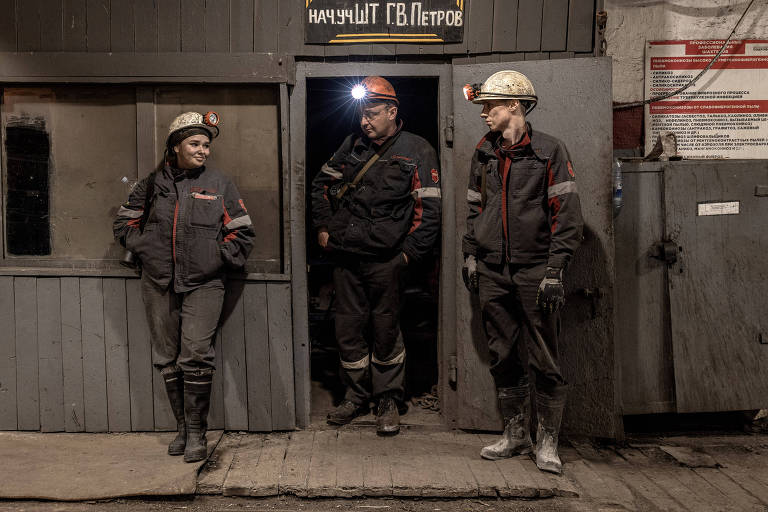 A imagem mostra três trabalhadores em uma mina, dois homens e uma mulher, todos usando capacetes de segurança e roupas de trabalho escuras. Eles estão em uma entrada de uma estrutura, com uma parede de madeira ao fundo e uma placa acima que parece ter texto em russo. O homem no centro usa um capacete laranja com uma lanterna. O ambiente é escuro, com iluminação suave. O chão é de madeira e há um equipamento visível ao fundo.