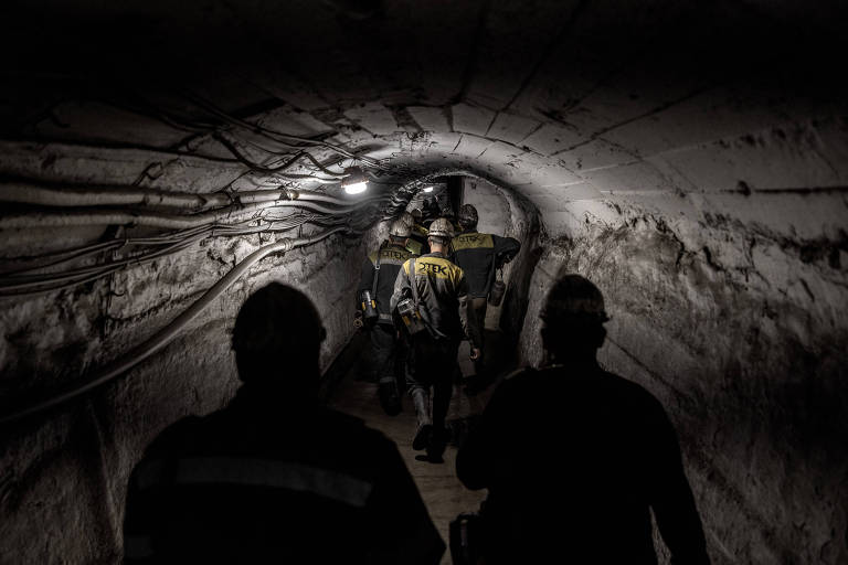 A imagem mostra um grupo de trabalhadores caminhando em um túnel de mina. O túnel é estreito, com paredes de concreto e fiação exposta no teto. Os trabalhadores usam capacetes e uniformes, alguns com luzes acesas. A iluminação é fraca, com uma luz artificial visível no teto, criando um ambiente escuro e industrial.