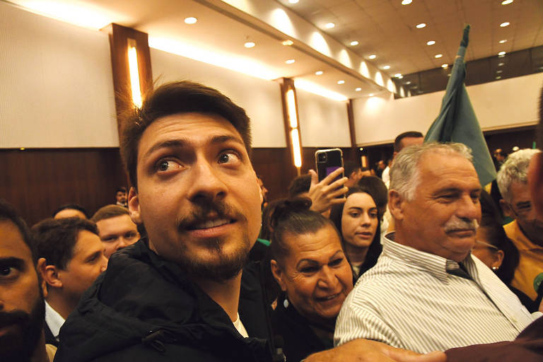 A imagem mostra uma multidão em um evento, com várias pessoas em um ambiente interno. Um homem com cabelo escuro e barba está em primeiro plano, olhando para a câmera. Ao seu redor, há outras pessoas, algumas sorrindo e outras olhando para o lado. Um homem mais velho, com cabelo grisalho, está à direita, usando uma camisa branca. Ao fundo, é possível ver uma bandeira verde e luzes do ambiente.