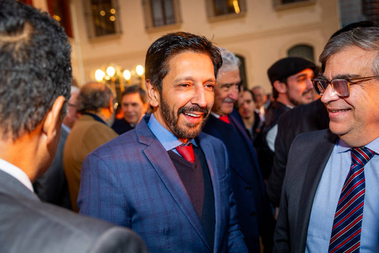 A imagem mostra um grupo de homens em um evento social. Um homem em destaque, com cabelo escuro e barba, está sorrindo e usando um terno azul com uma gravata vermelha. Ao seu lado, outro homem também sorri, usando óculos e um terno escuro. O fundo é composto por outras pessoas e um ambiente iluminado, sugerindo um evento festivo.