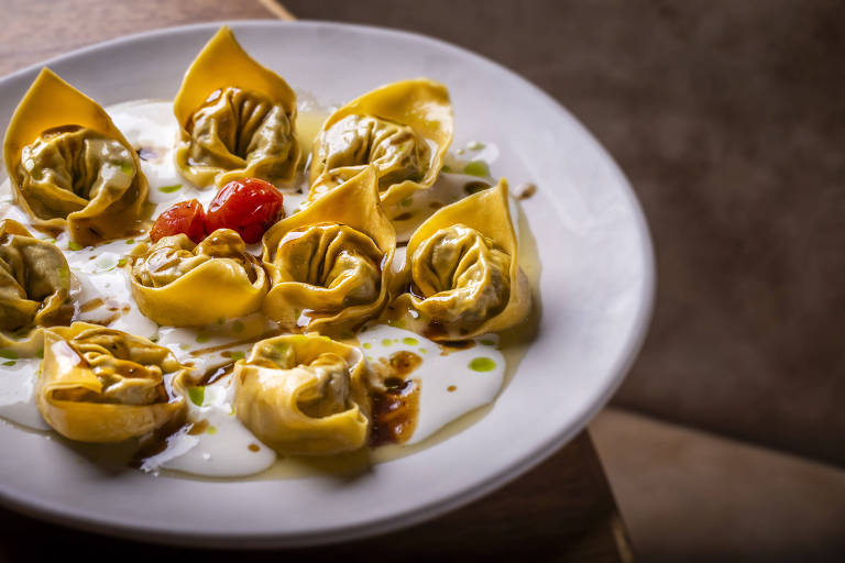 Um prato redondo com tortelli amarelo, recheado, dispostos em círculo. No centro, há três tomates-cereja vermelhos. O prato é decorado com um molho branco e um fio de azeite.