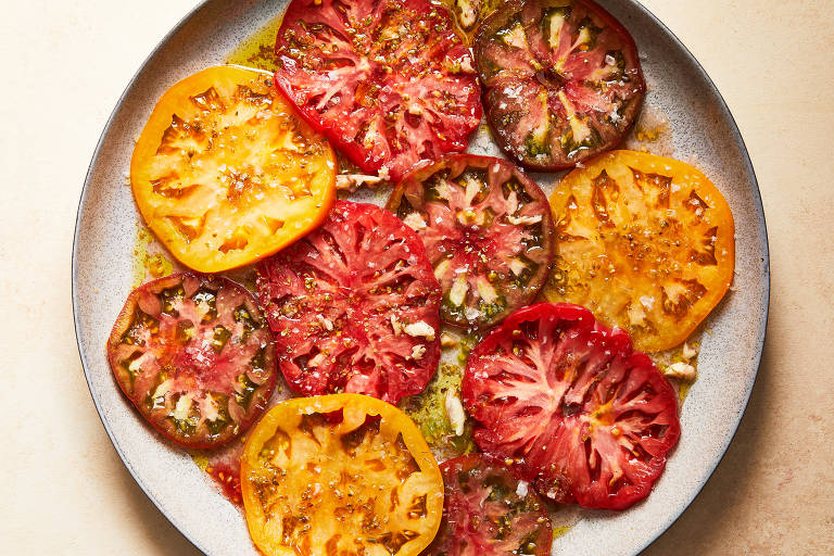 Um prato redondo com várias fatias de tomate em diferentes cores, incluindo amarelo, vermelho e marrom. As fatias estão dispostas de forma sobreposta e cobertas com um molho brilhante, possivelmente de azeite, e polvilhadas com sal. O fundo do prato é de cor clara, contrastando com as cores vibrantes dos tomates.