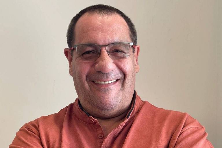 Homem de meia-idade está sorrindo e posando para a foto. Ele usa óculos e uma camisa laranja. Seus braços estão cruzados sobre o peito, e ele está em um ambiente com parede clara ao fundo