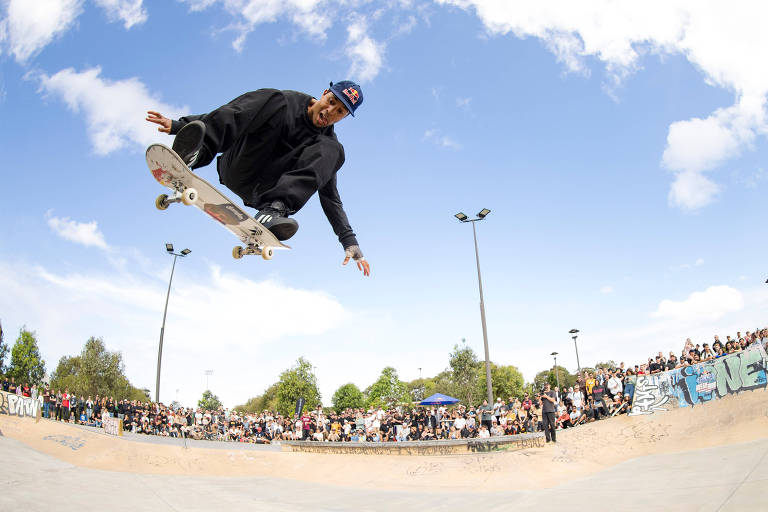 Um skatista está realizando um salto no ar em um parque de skate, com o céu azul e algumas nuvens ao fundo. Há uma multidão de pessoas assistindo ao redor da pista, e o skatista está vestido com roupas escuras e um boné. A cena captura a energia e a emoção do esporte.