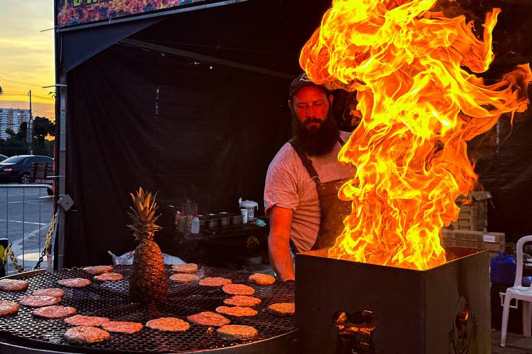 A imagem mostra um homem com barba e boné, cozinhando em uma grelha com chamas altas. Ele está em um ambiente de food truck, com hambúrgueres grelhando em uma grade circular. No centro da grelha, há um abacaxi. Ao fundo, é possível ver um pôr do sol e alguns carros estacionados.