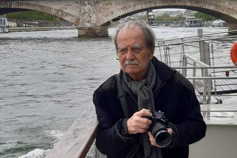 Um homem calvo, com cabelo e bigode grisalhos, está ao lado de um parapeito de madeira ao ar livre, segurando uma câmera. Ele usa um casaco escuro, e há um rio ao fundo. Há uma ponte visível atrás dele e o céu está nublado.