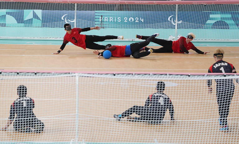 A imagem mostra uma partida de goalball, com jogadores caindo no chão em uma quadra. Dois jogadores estão deitados, um usando uma bola azul, enquanto outros dois estão em movimento. No fundo, há uma rede e o logotipo dos Jogos Paralímpicos de Paris 2024.