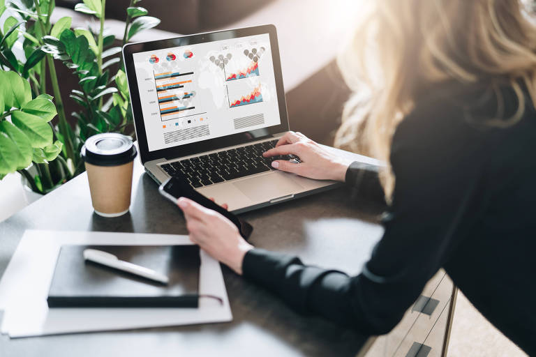 A imagem mostra uma mulher sentada em uma mesa, usando um laptop. A tela do laptop exibe gráficos e dados. A pessoa está segurando um celular com a mão direita e tem um copo de café ao lado, em uma mesa de madeira. Há um caderno e uma caneta sobre a mesa, além de plantas ao fundo.
