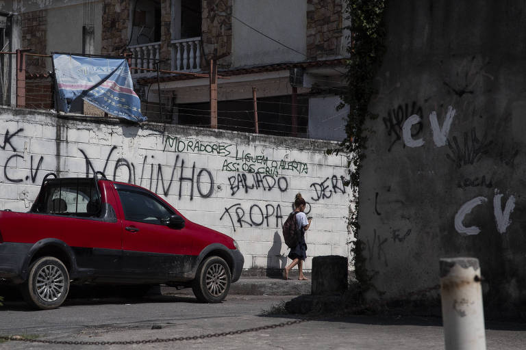 A imagem mostra uma rua urbana com um carro vermelho estacionado à esquerda. Ao fundo, há uma parede coberta de grafites em tinta preta, com palavras e frases escritas. Uma pessoa está de costas, próxima à parede, aparentemente observando os grafites. O ambiente é ensolarado e há uma estrutura de edifício visível ao fundo.
